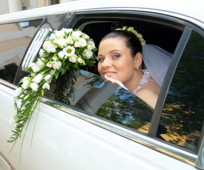 Braut mit Blumenstrauß schaut aus einer weißen Stretchlimousine